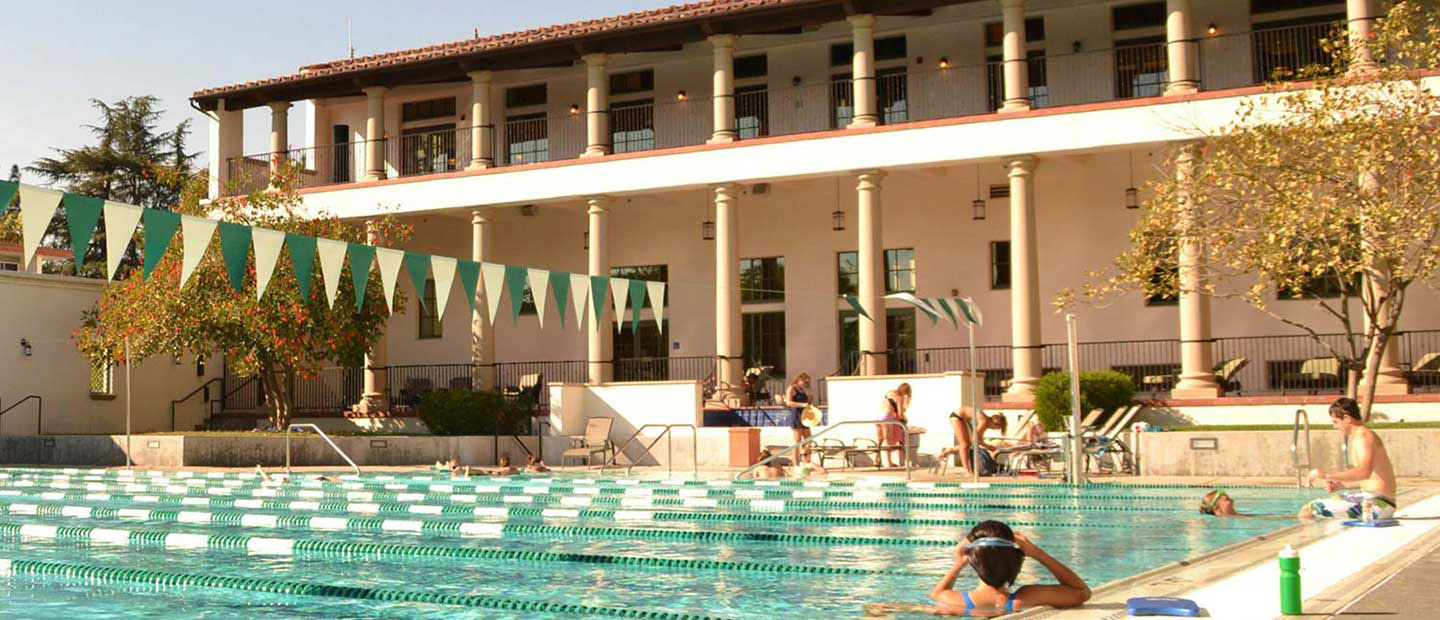 A pool with swimmers resting on the side. Behind is a two-story building with a columned terrace on each level.