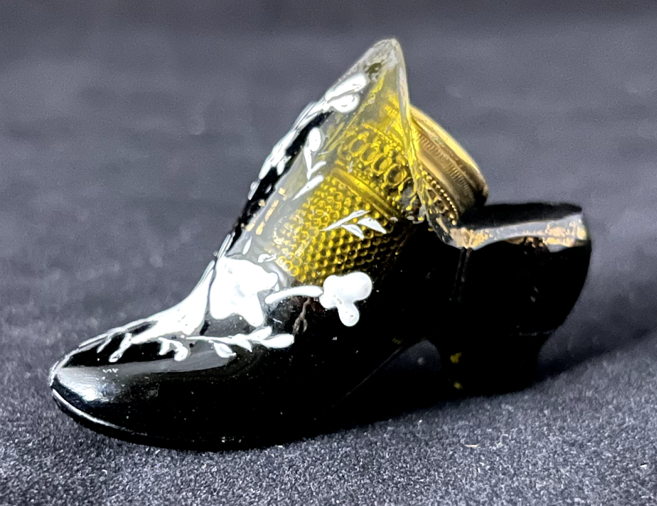 A decorated brass thimble in an enameled dark green glass holder in the shape of a shoe.
