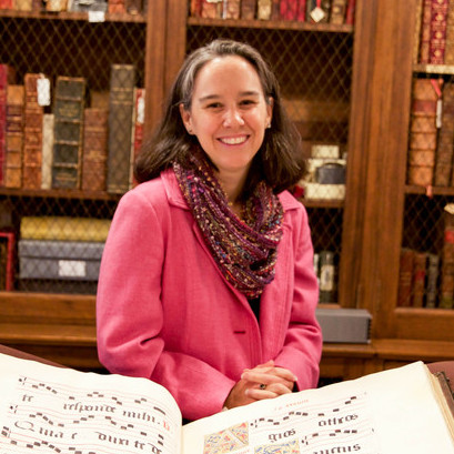 White woman with brown hair and a pink coat in front of a book