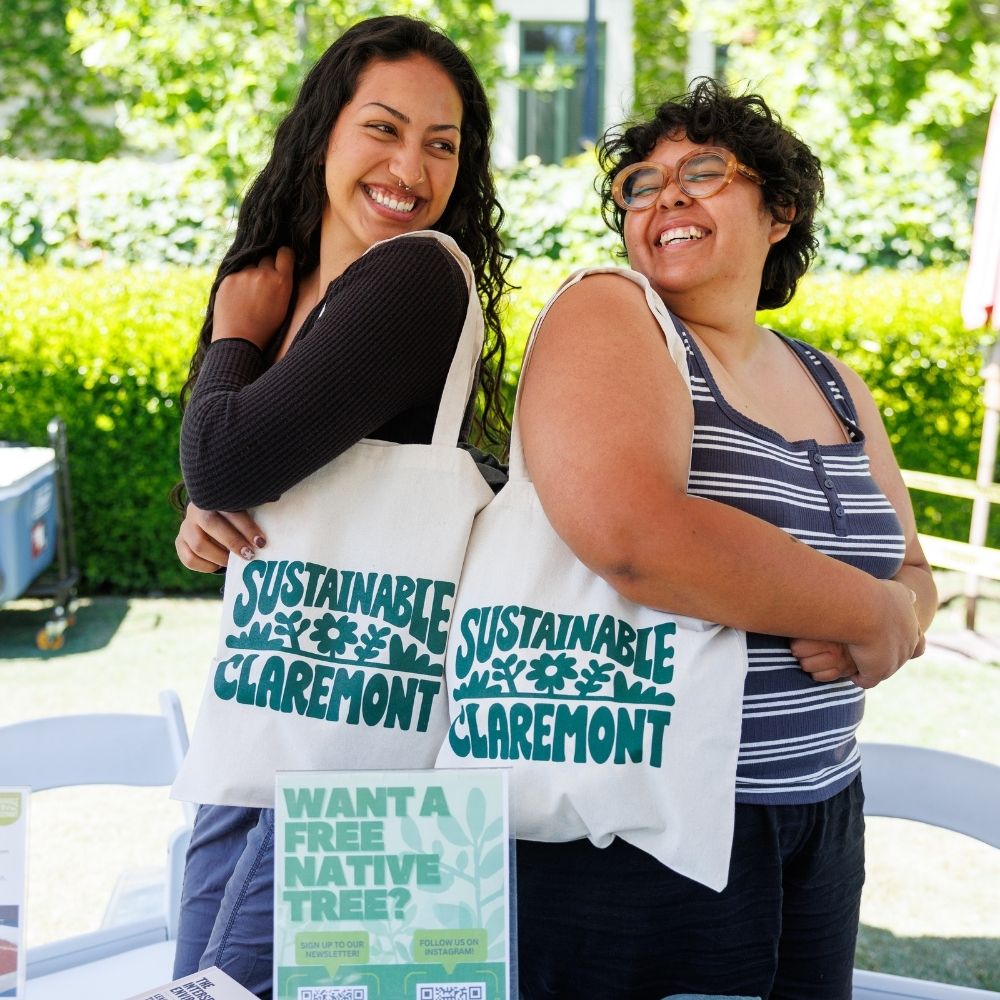 Two women wearing tote bags stand back-to-back and smile at each other