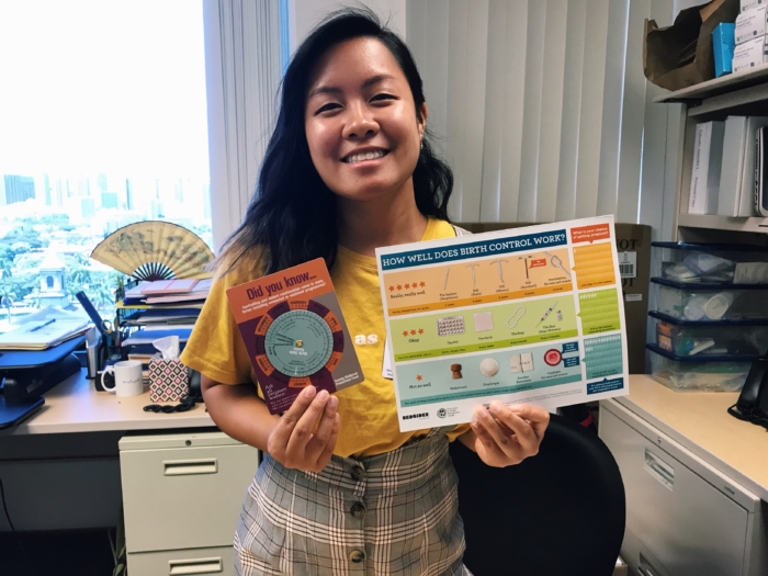 A young Hawaiian woman in a yellow t-shirt and plaid coveralls holding up informational cards about birth control.