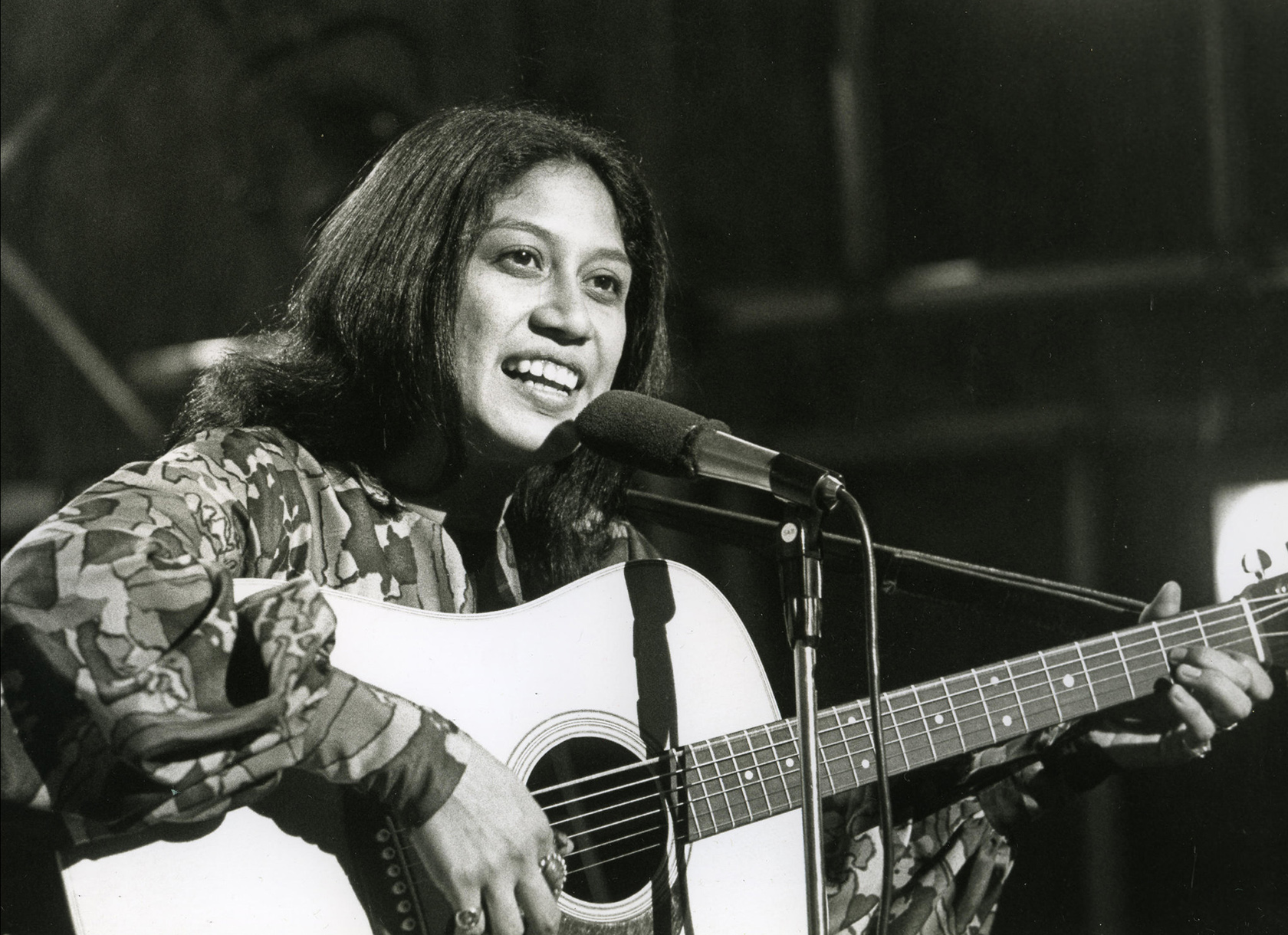 A black and white photo of a woman playing a guitar and singing into a microphone.