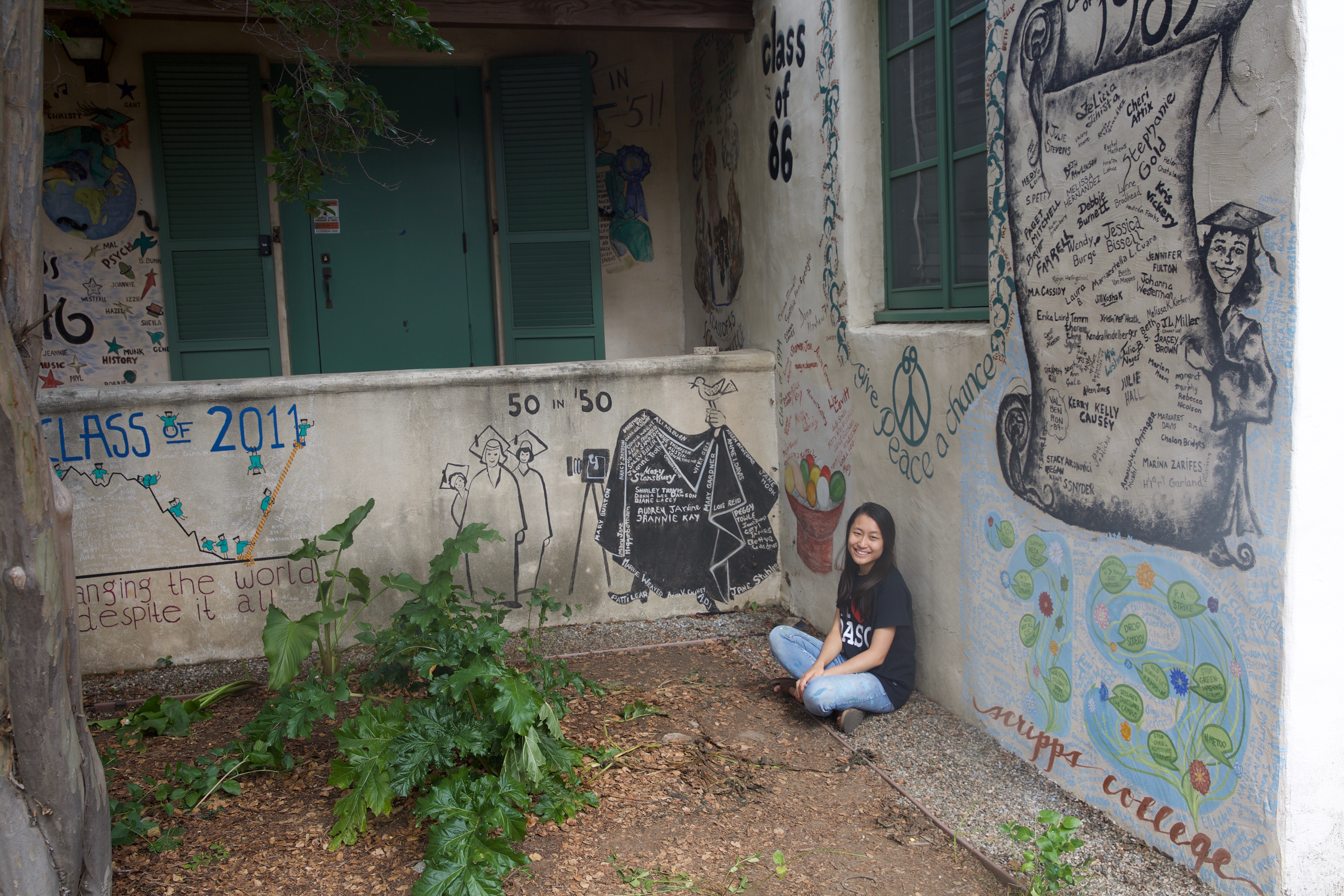 A young Asian woman sitting in a courtyard against a wall covered in graffiti art.