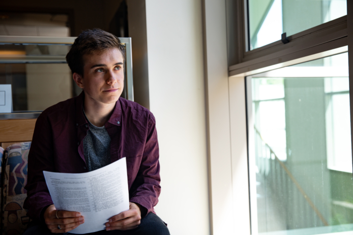 A young man with short brown hair and wearing a red shirt sitting indoors with a paper in his hands and looking out a window.