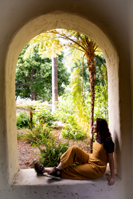 A young woman with long brown hair and tan overalls sitting on a window sill looking outside.