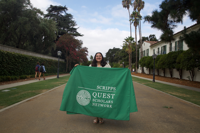 A young woman with curly brown hair and glasses holding a banner saying 'Scripps Quest Scholars Network'.