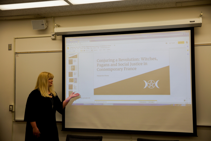 A young white woman in formal clothing presenting a powerpoint at the front of a classroom.