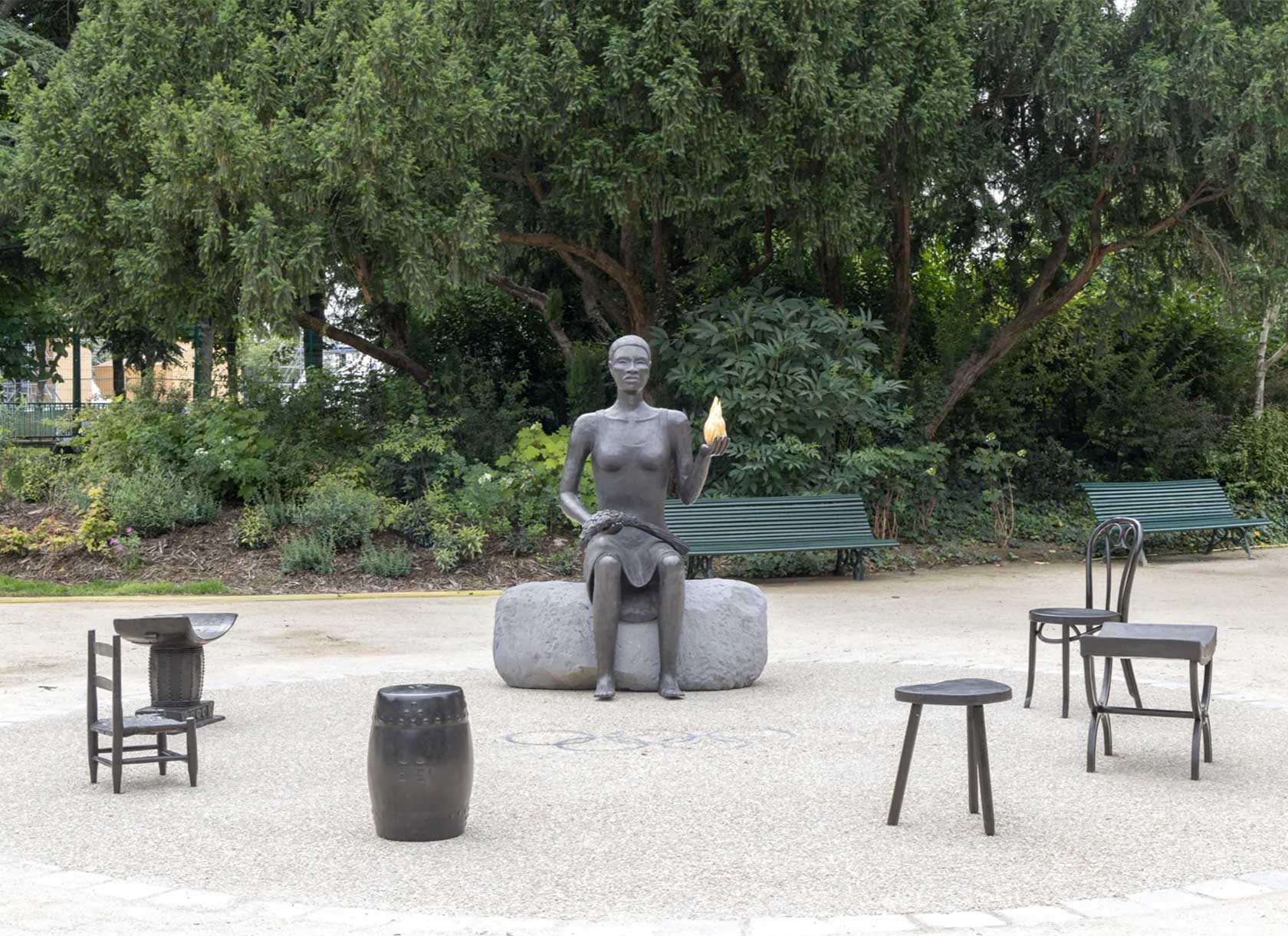A bronze sculpture of a Black woman is seated in a garden setting. The sculpture is surrounded by six chairs of different designs.