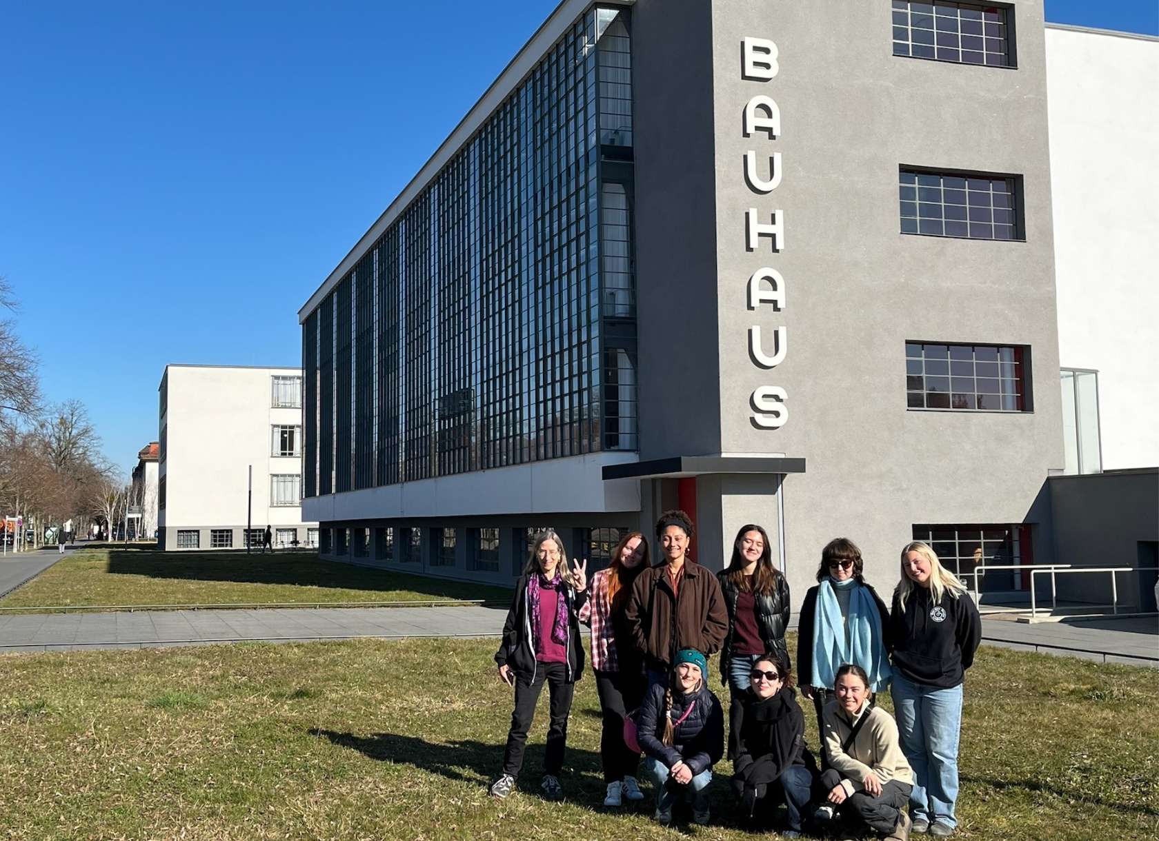students gather on a lawn in front of a building with a large BAUHAUS sign running alongside its facade