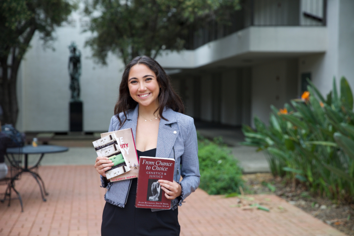 A young woman in formal clothing smiling and holding up books in front of a classroom building.