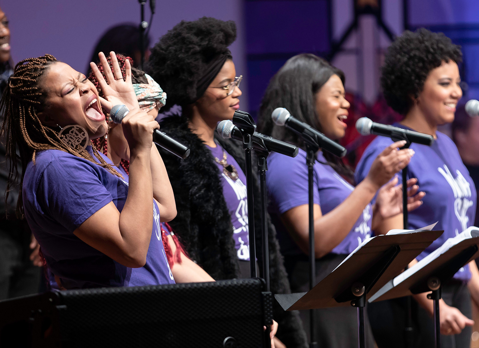 Four young black women wearing purple T-shirts and singing into microphones.