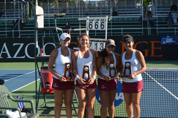 Four young women in sports uniforms posing with awards on a tennis court.
