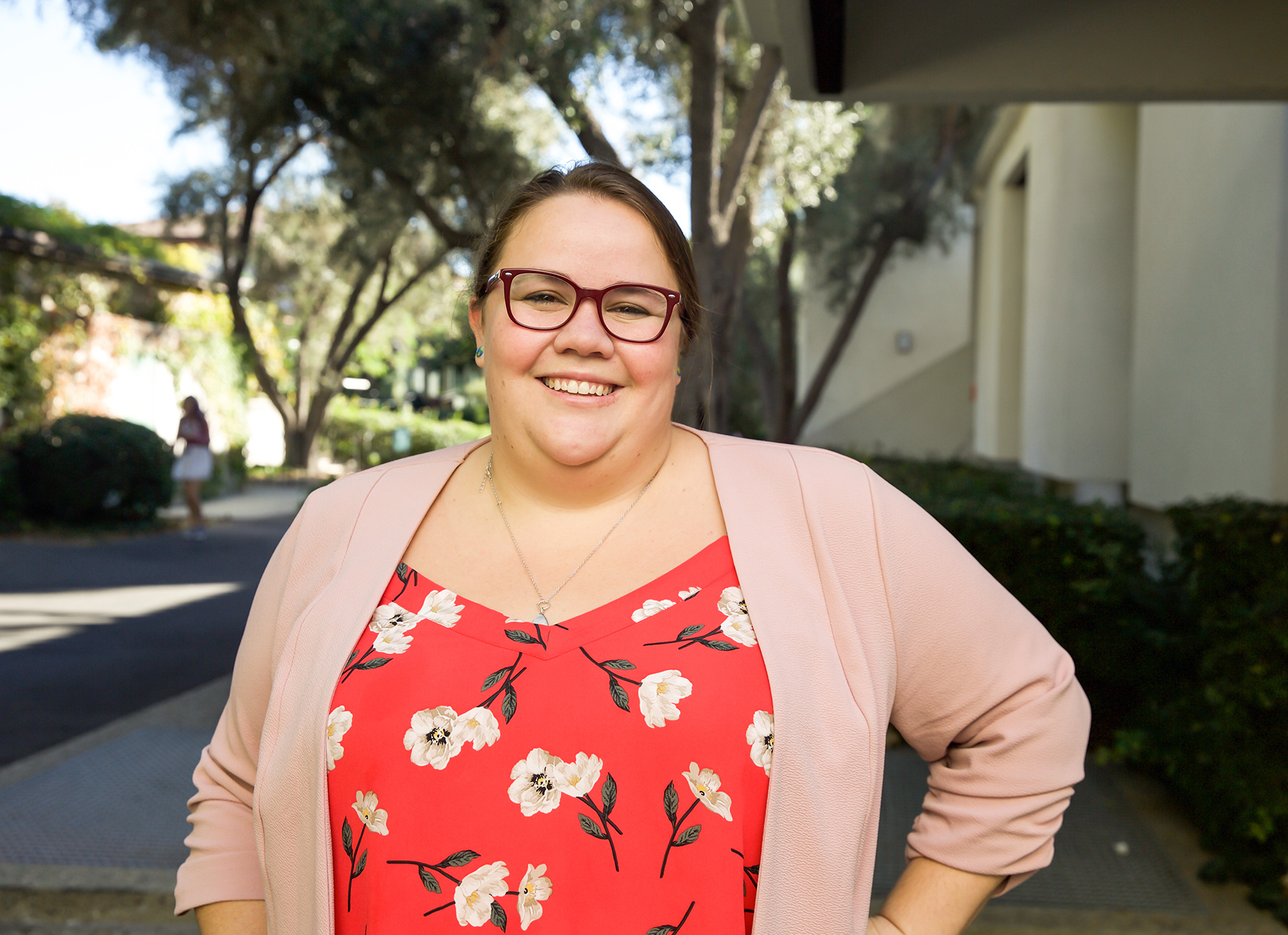 A white woman with brown hair and glasses wearing a pink sweater and red floral shirt smiling with one hand on her hip.