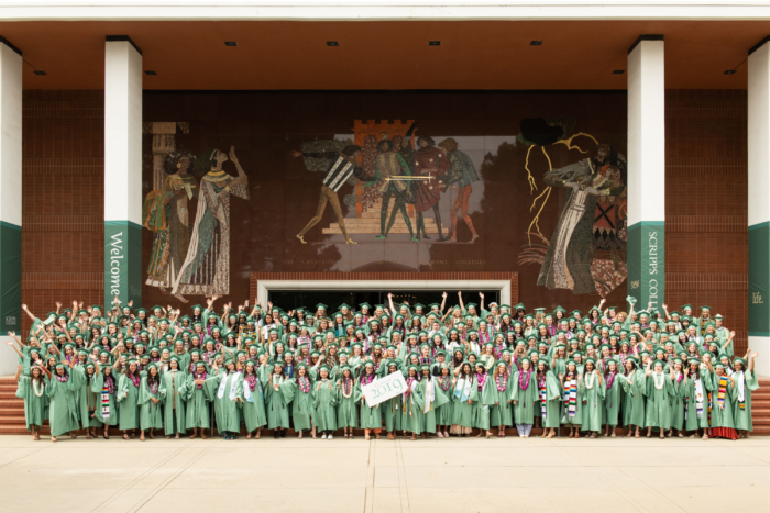 A crowd of young people in graduation caps and gowns standing in front of a theater with a sign saying 'Class of 2019'.