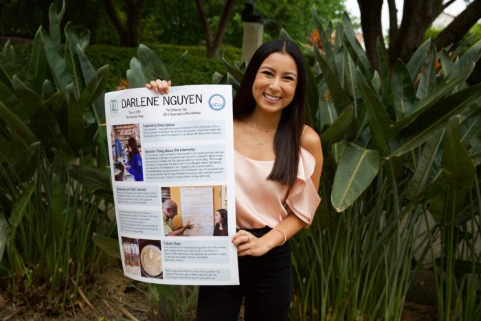 A young Asian woman in formal clothing smiling and holding up a poster outside.