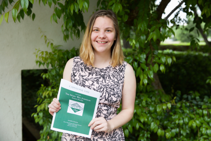 A young white woman in a formal dress holding up a flyer for 'Capstone Day' outside.