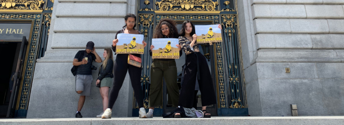 Three young women posing in front of a city hall building holding signs that say 'Shut Down 850'.