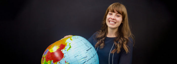 A young woman with long brown hair smiling and wearing a professional blue sweater holding an inflatable globe.