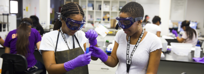 Two young African American women wearing goggles and gloves white looking at a test tube in a science lab.