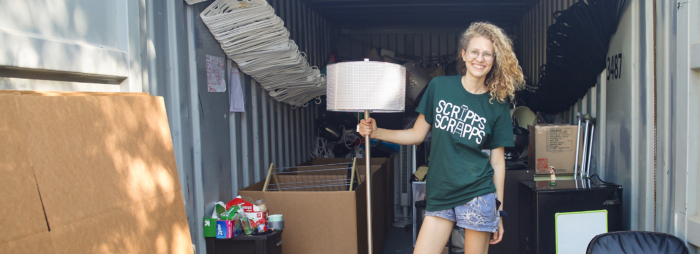 A young woman with blond curly hair and glasses wearing denim shorts and a t-shirt saying 'Scripps Scrapps" smiling and holding a lamp.