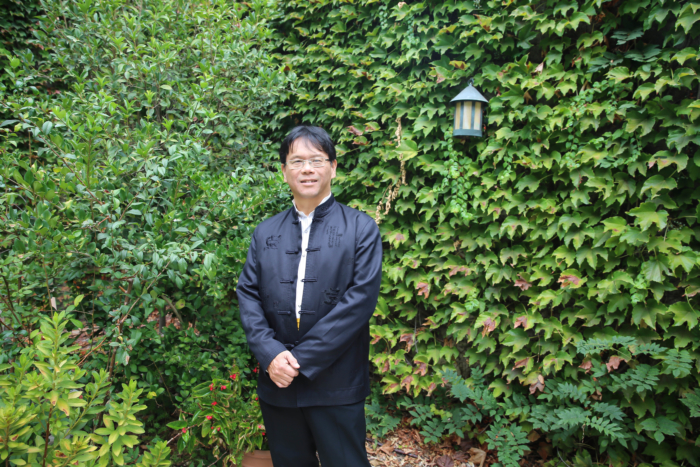 An Asian man wearing glasses and black traditional clothing smiling in front of a nature background.