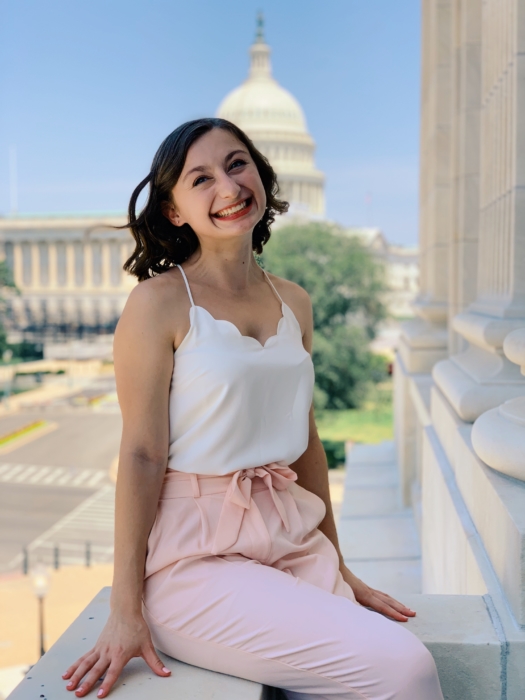 A young white woman with short brown hair wearing a white tank top and pink pants sitting on a ledge with the Capitol Building in the background.