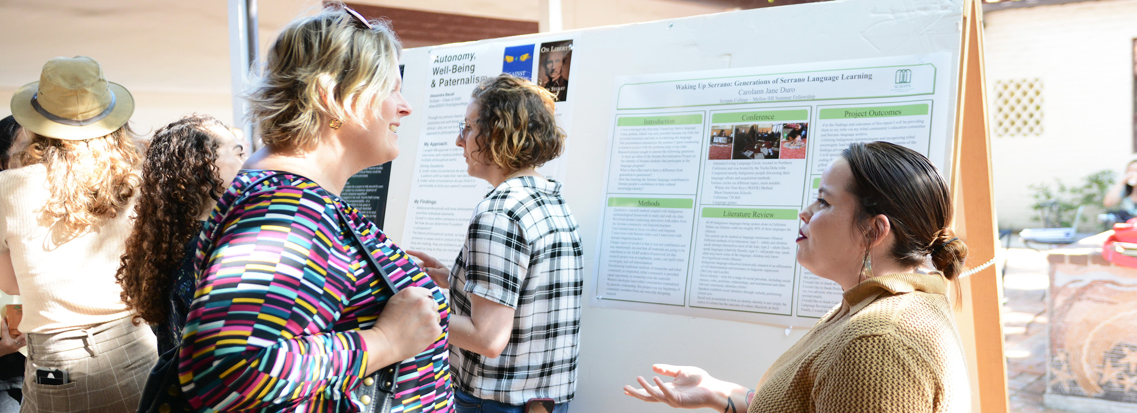 A woman with brown hair in a ponytail talking to a smiling blond woman outside in front of a research poster.