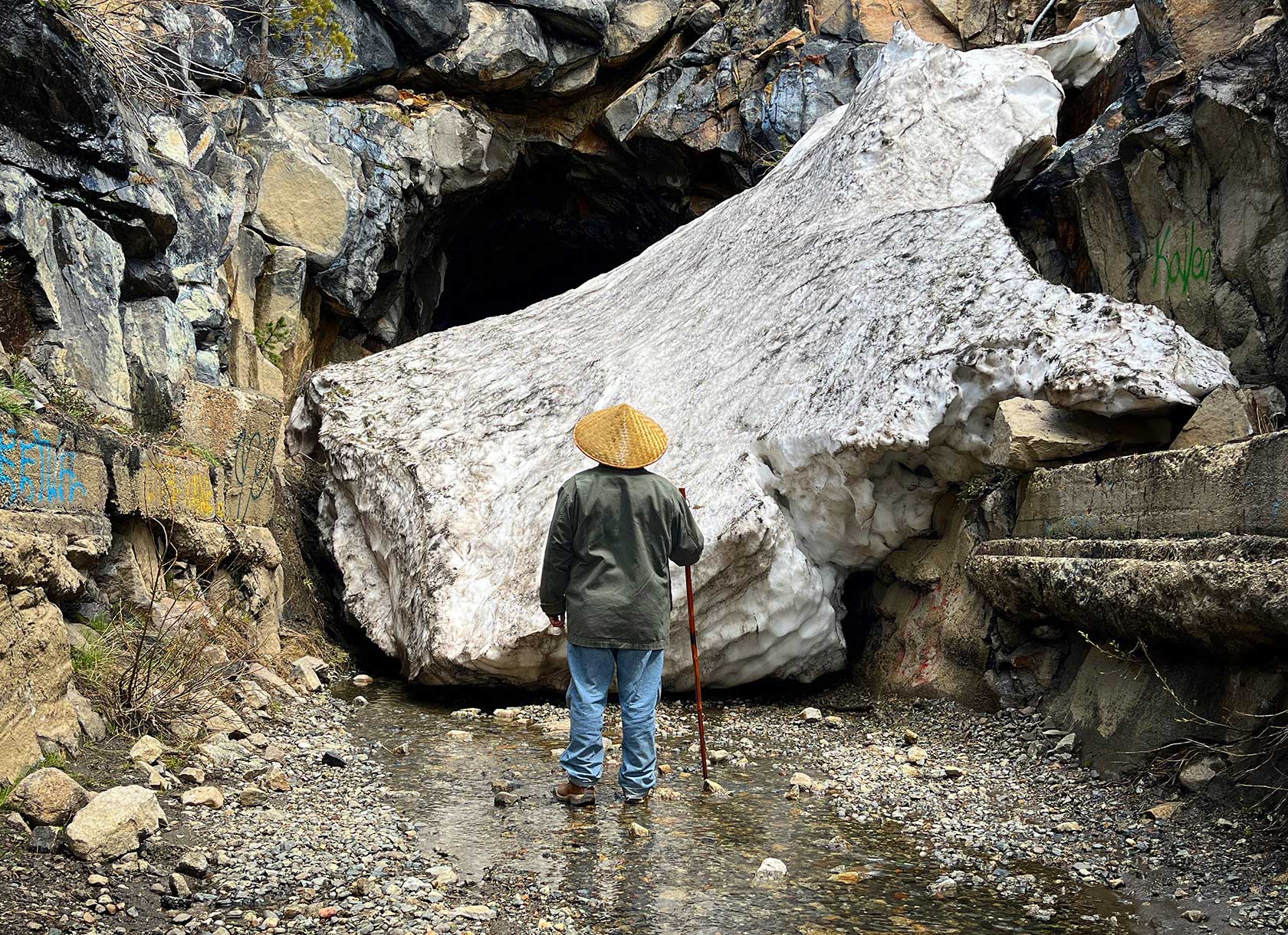 A man in a hat stands in a river, looking toward a rock face