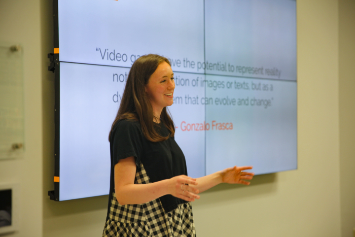 A young white woman in formal clothing giving a presentation at the front of a room.