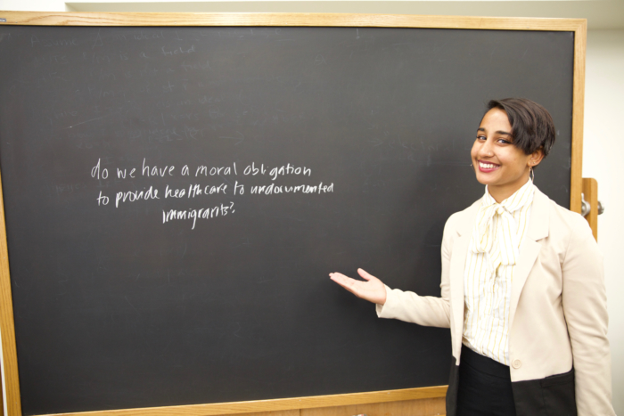 A young South Asian woman in formal clothing standing at a blackboard.