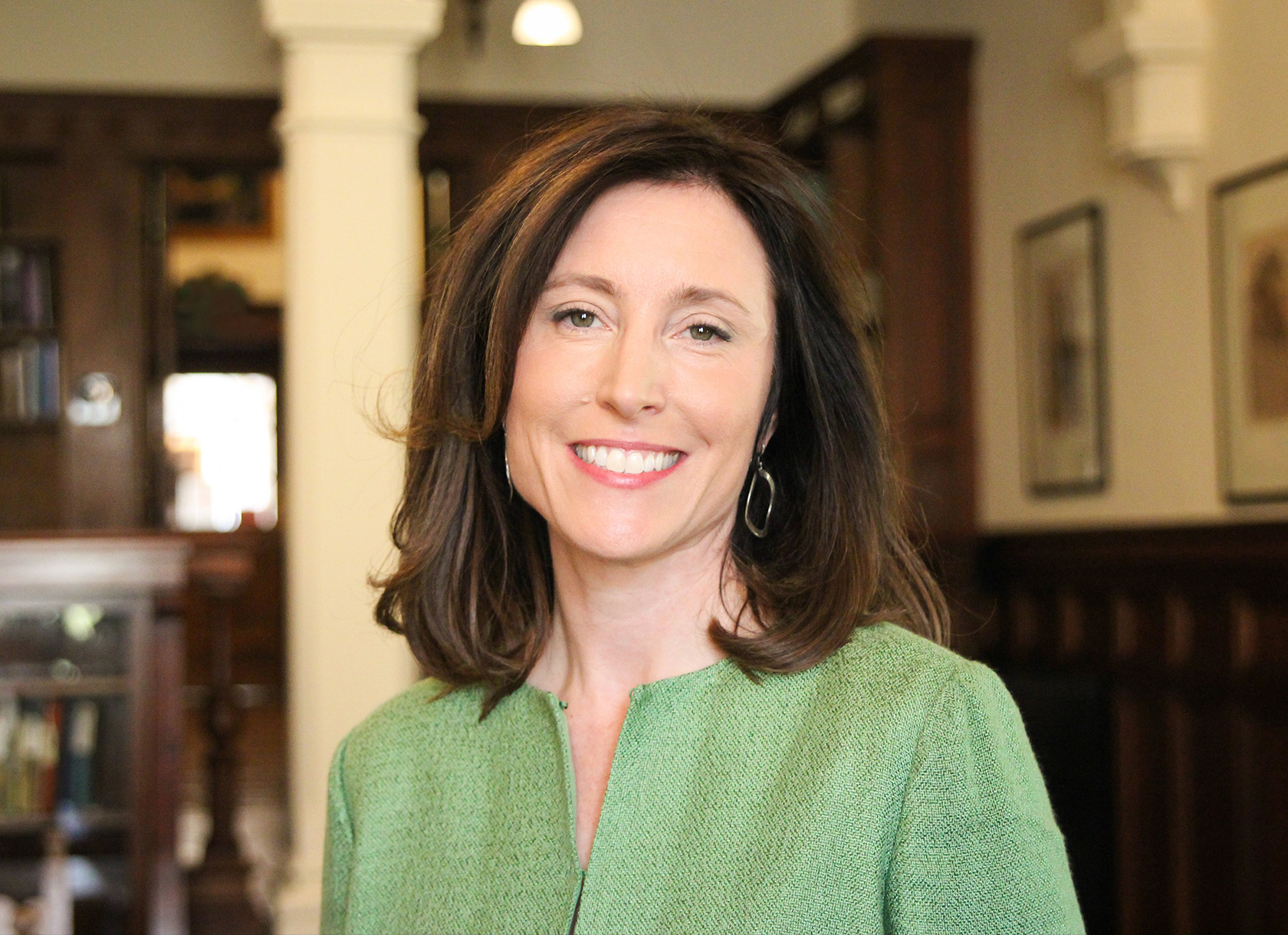 A headshot of a smiling white woman with shoulder-length brown hair wearing a green blouse and standing in a library.