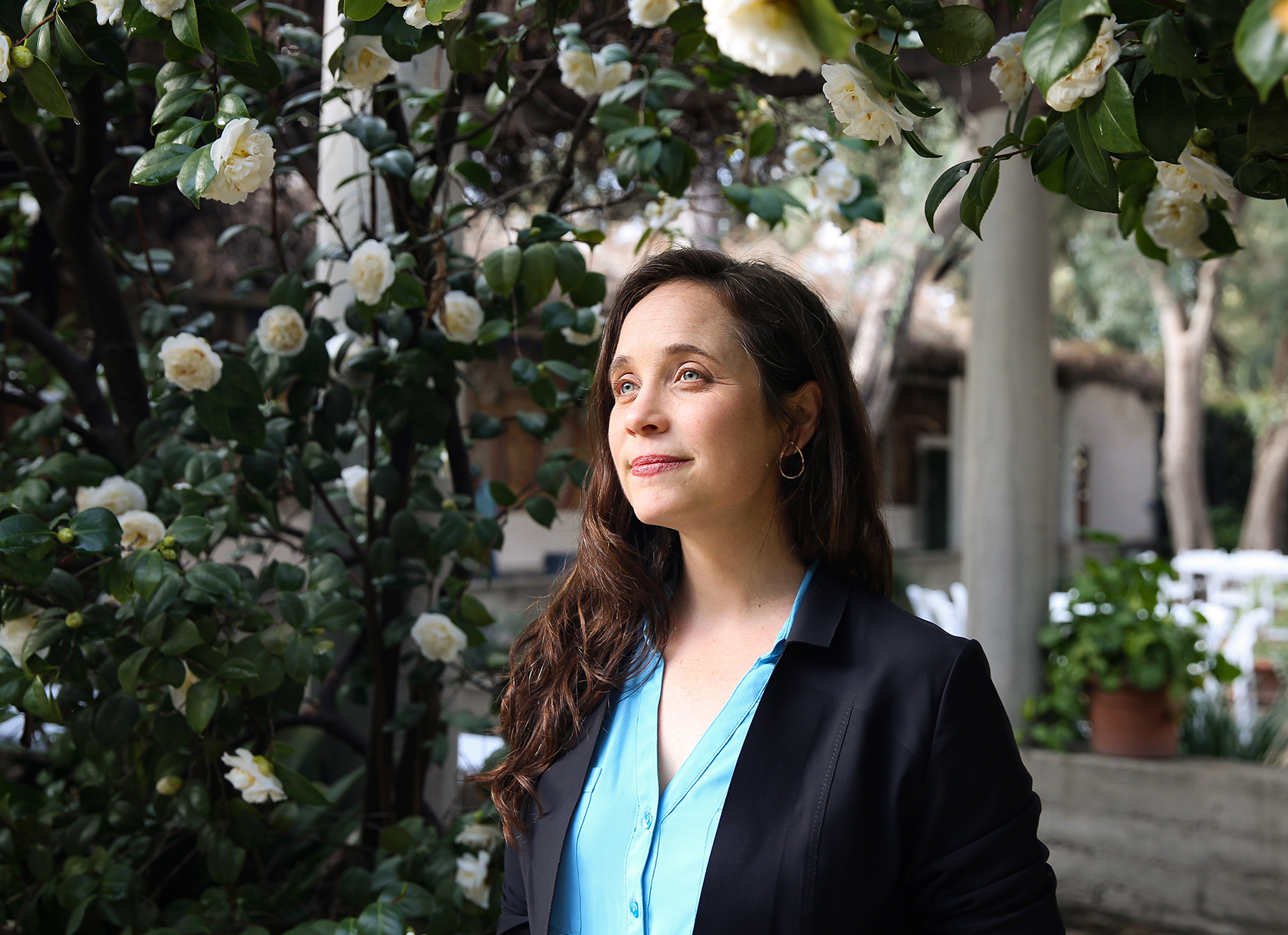 A woman in professional clothes with brown hair and pale eyes smiling in a garden.