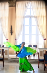 A woman in brightly colored green and red traditional clothing dancing in a sunlit room.