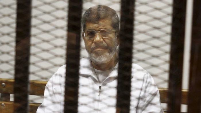 A Middle Eastern man with gray hair, a beard, glasses, and a white jacket sitting on a bench on the other side of a gate.