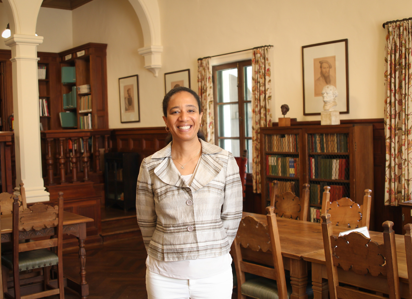 A smiling black woman with short black hair and wearing a white and gray pantsuit standing in a library. 