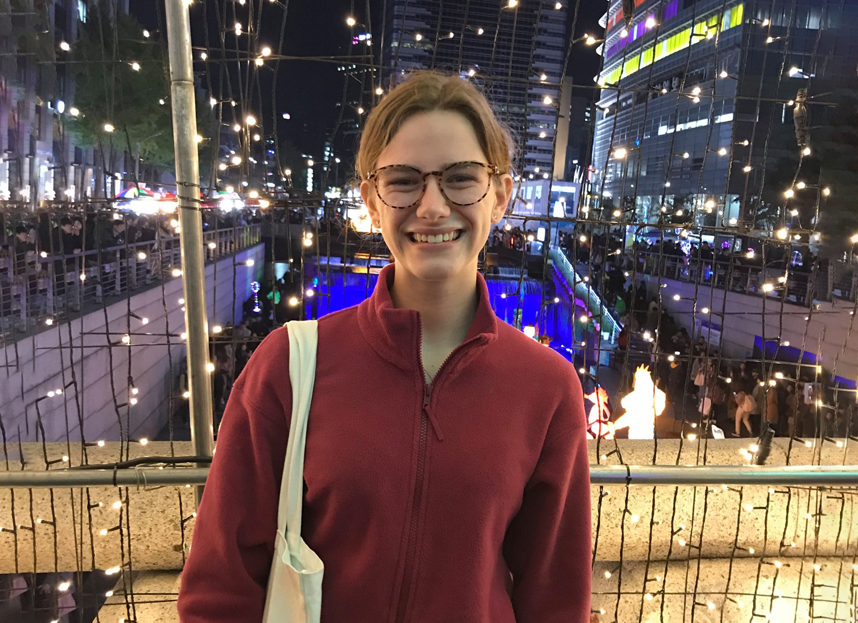 A young white woman with brown hair wearing glasses and a red jacket and smiling in front of a city background.