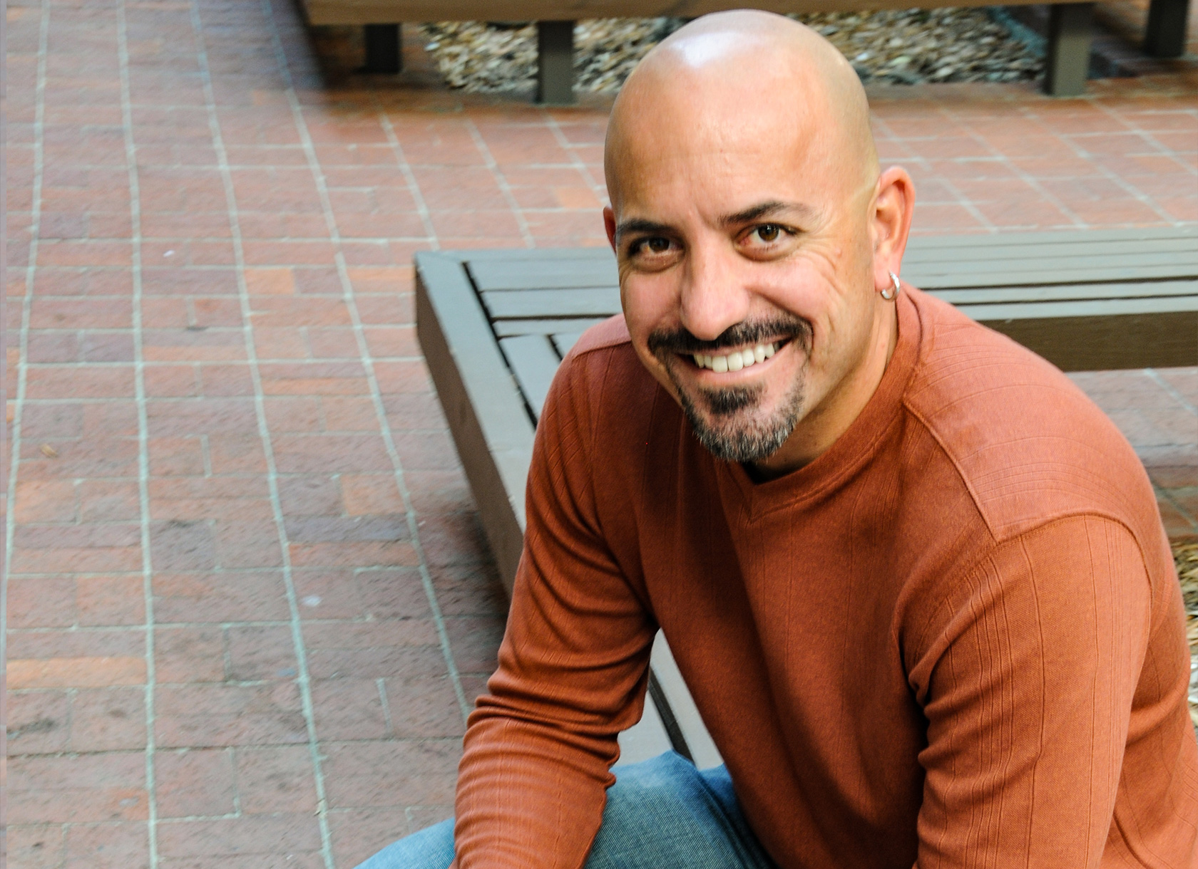 A bald, bearded man in an orange sweater and jeans smiles at the camera from a seated position