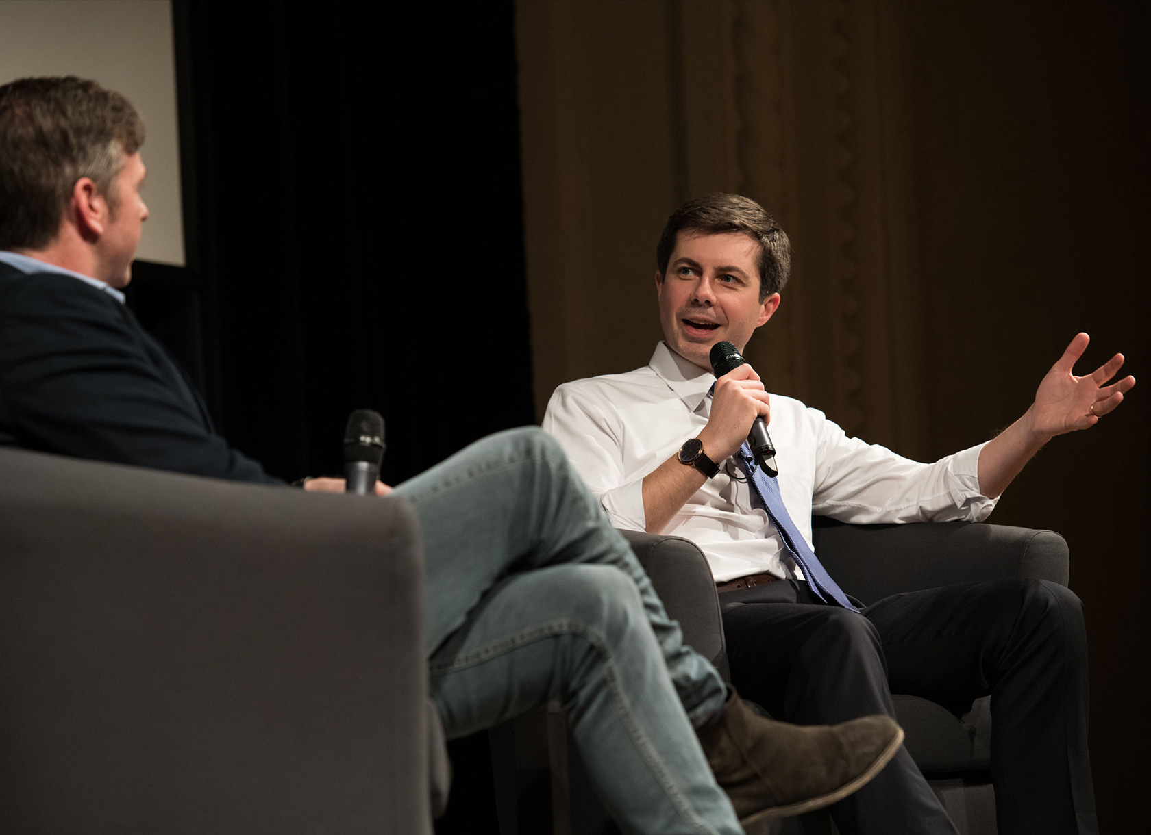 Pete Buttigieg sitting onstage and talking into a microphone with another white man.