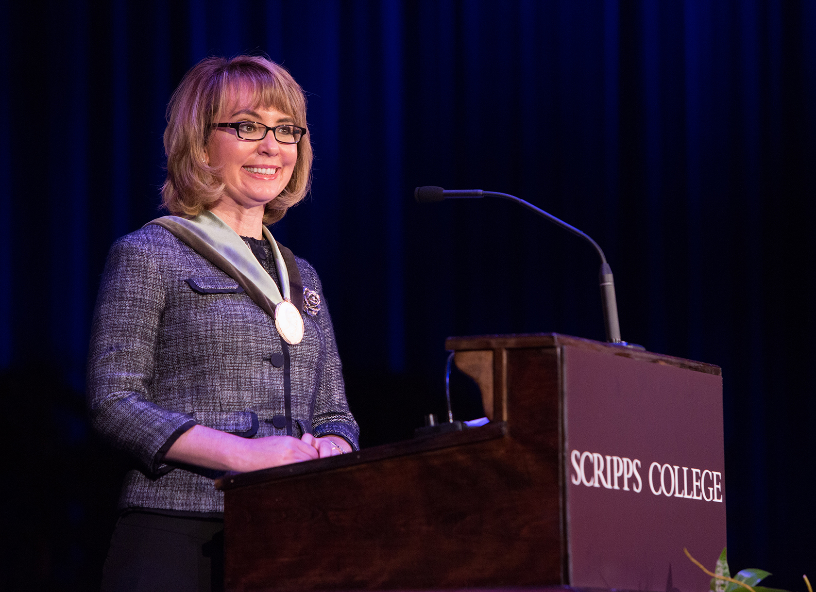 A white woman with short blonde hair and glasses stands behind a Scripps College podium