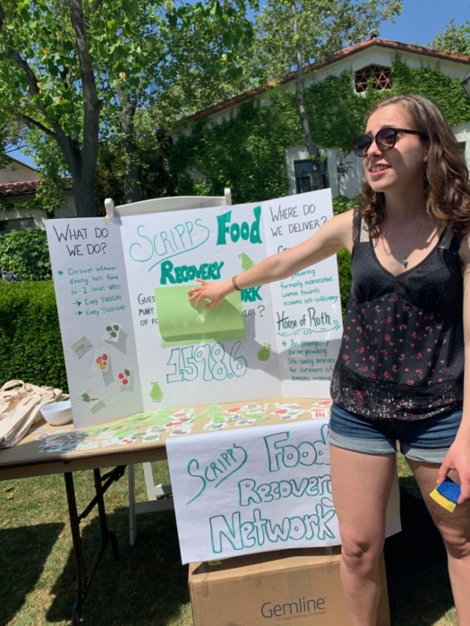 A young white woman in shorts and a tank top standing outside in front of a table advertising the Scripps Food Recovery Network.