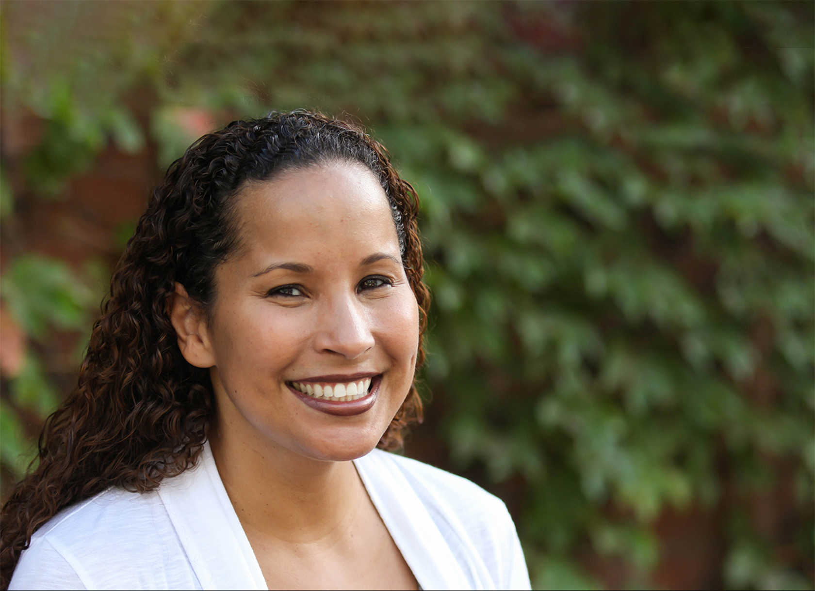 A headshot of a black woman with long, curly hair and a white blazer smiling at the camera.