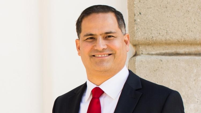 A white man with salt and pepper hair wearing a black suit and red tie and smiling at the camera.