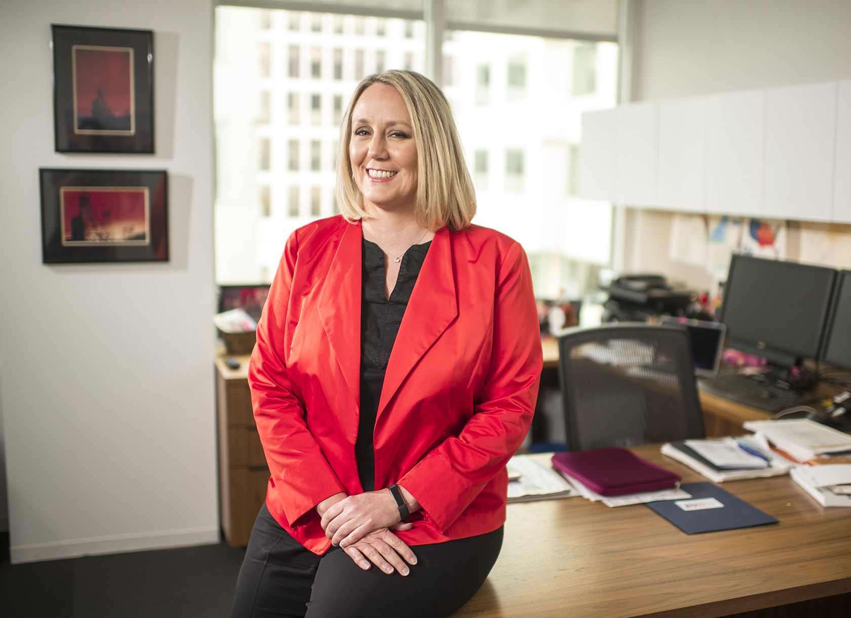 A white woman with short blond hair and a red blazer sitting at the edge of a desk and smiling.