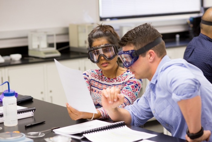 Two people wearing lab goggles and looking at papers while working at a lab table.