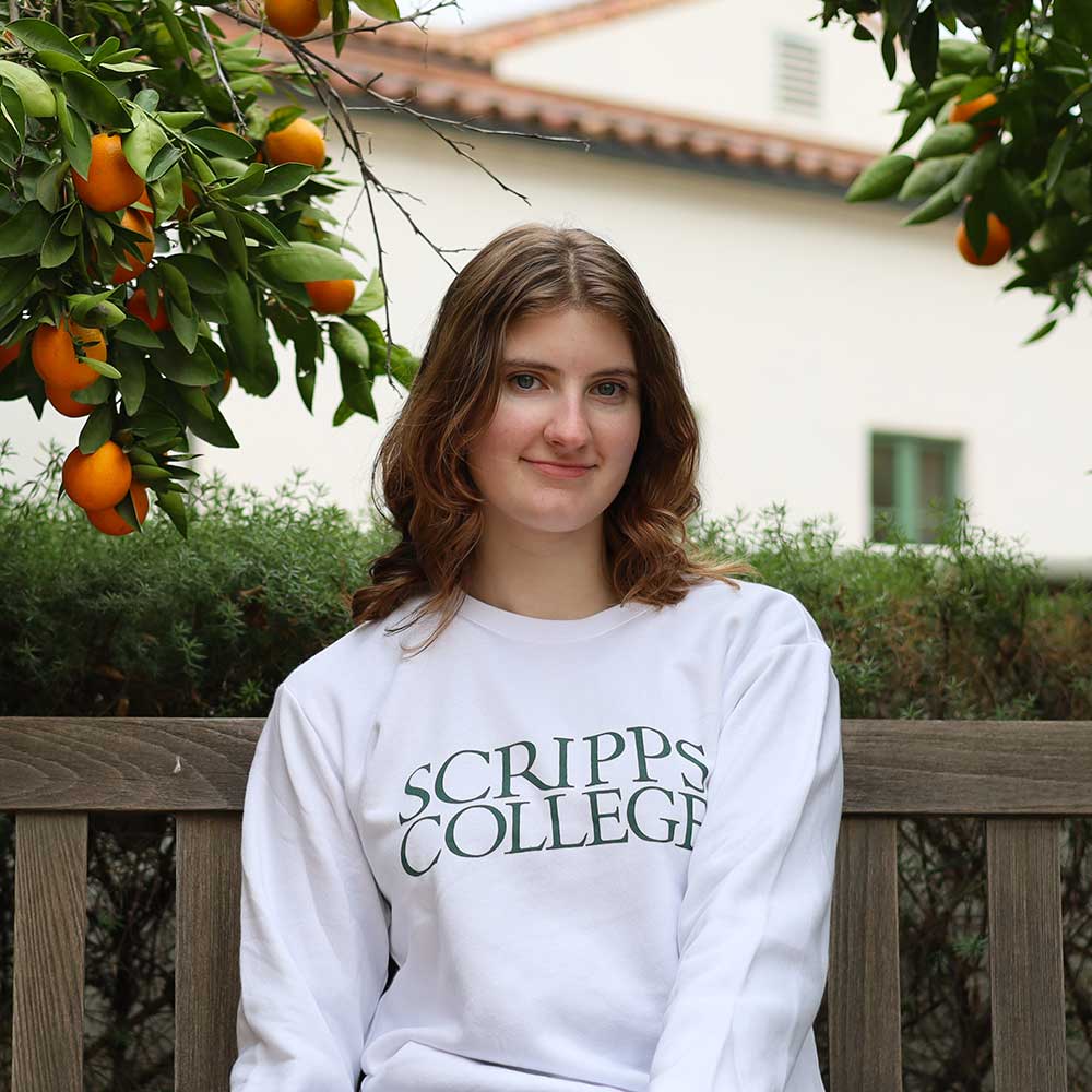 Sutdent on a bench near and orange tree in a white Scripps branded sweatshirt
