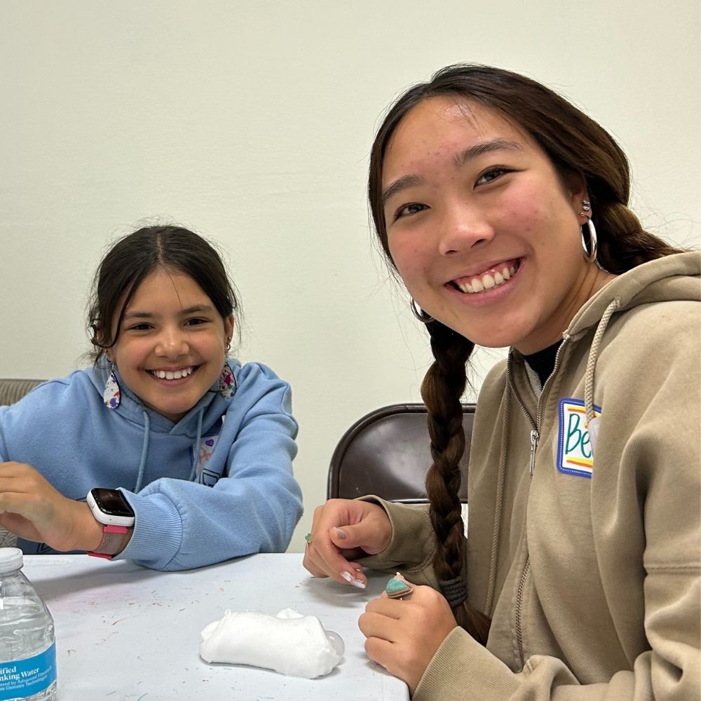 A young asian woman with her hair in two braids smiles while crafting next to a young child